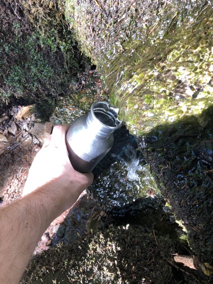 A hand collecting water from a natural mountain spring in a thermos