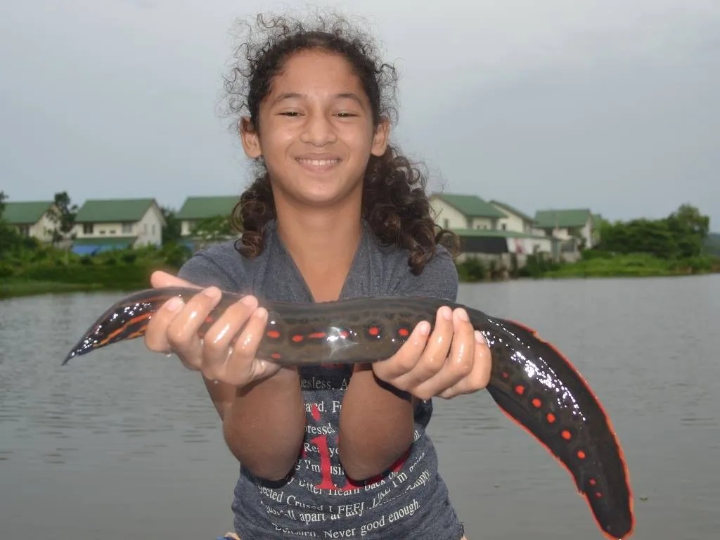 A young woman smiling as she is holding a fire eel across both her hands that she managed to catch in the river behind her that has a few homes at its edge.